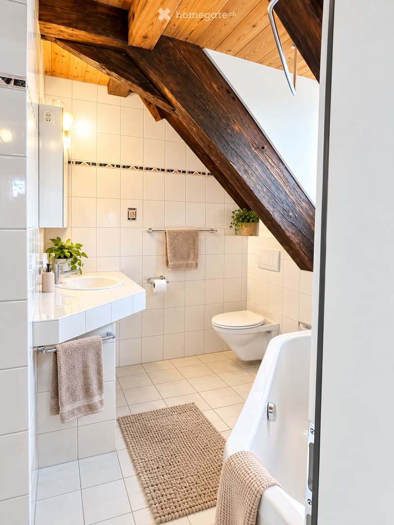 Modern bathroom with white tiles, a bathtub, toilet, sink, and decorative plants. Features a wooden slanted ceiling and warm lighting.