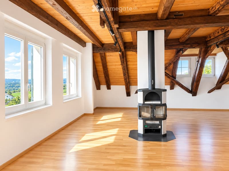 Bright, spacious room with exposed wooden beams and a modern wood-burning stove. Large windows offer a scenic view of the landscape.