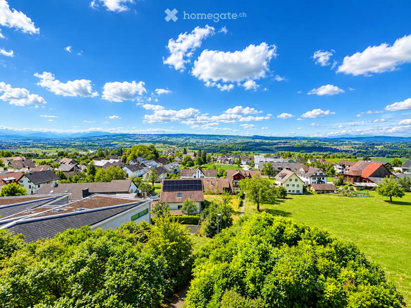 A panoramic view of a residential area with multiple houses, green lawns, trees, and a clear blue sky with scattered clouds. The landscape includes rolling hills and distant mountains.