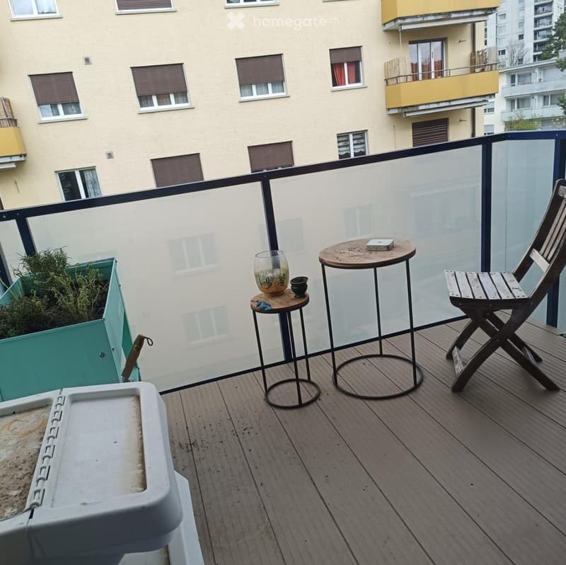 Balcony with wooden decking, glass railing, small round table, folding chair, and decorative items.