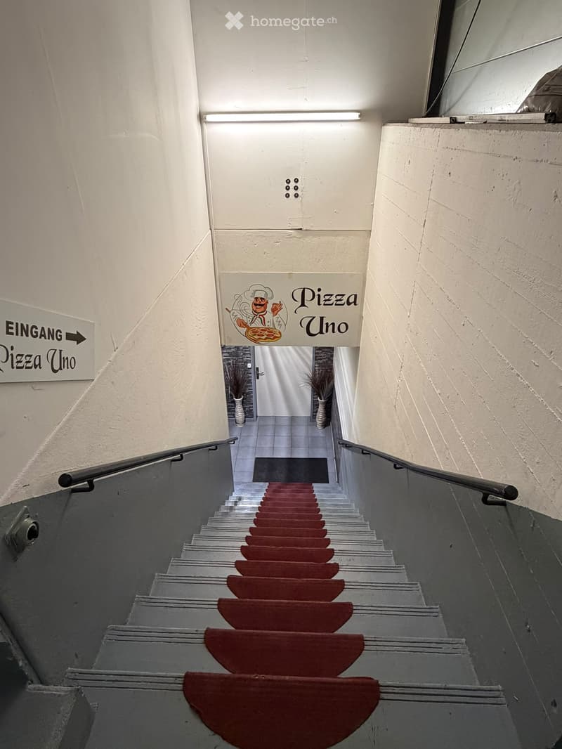 Staircase with red carpeted steps and gray risers, flanked by white walls and metal railings. Signage for "Pizza Uno" is visible at the top of the stairs.