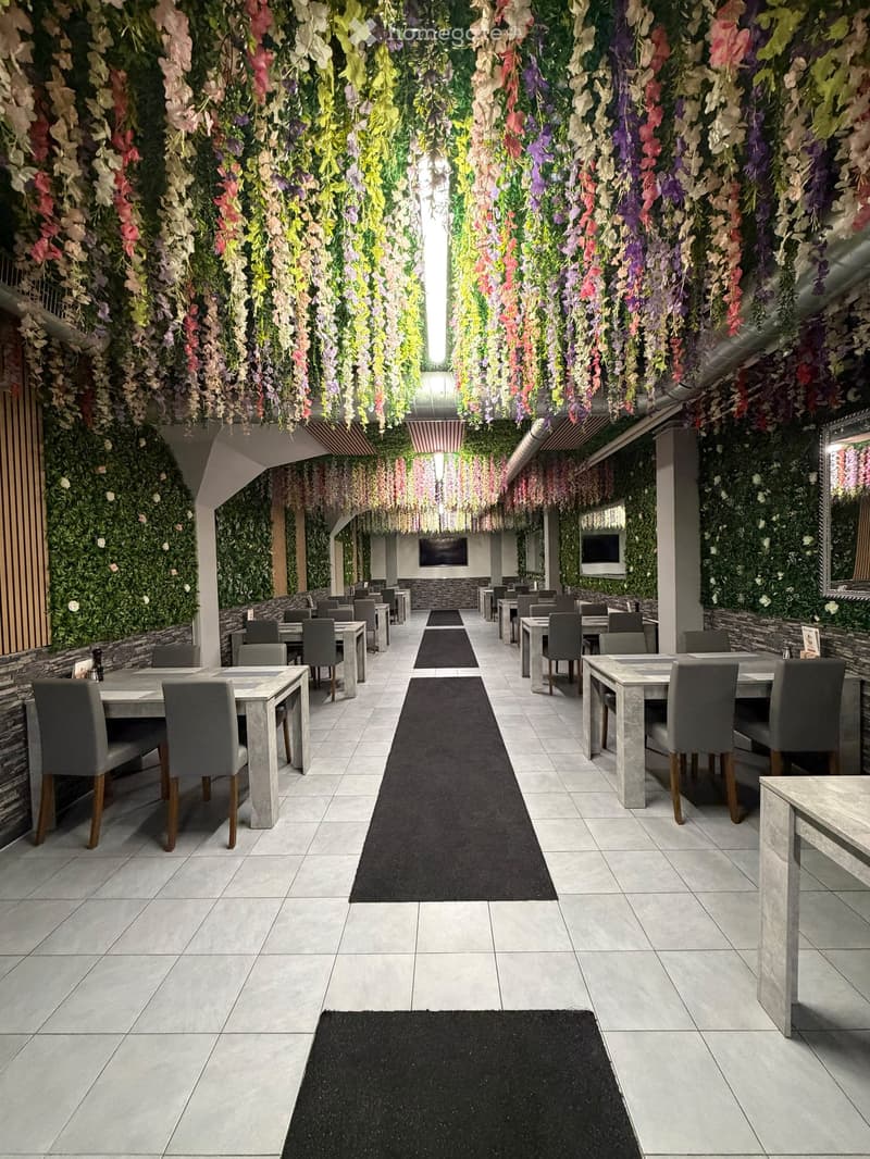 A modern restaurant with a long central aisle, multiple tables and chairs, and a floral vertical garden on the walls.