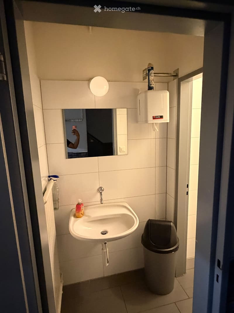Small bathroom with a white sink, mirror, hand dryer, soap dispenser, and a trash can. The walls are tiled in white, and the floor is tiled in a light color.