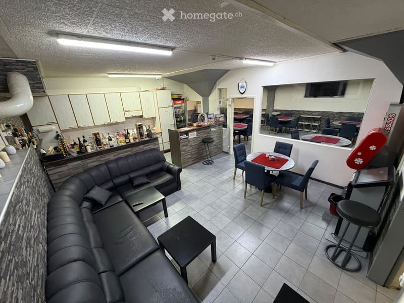 Modern furnished room with a black leather sofa, coffee table, bar area with stools, dining table with chairs, and a refrigerator in the background.