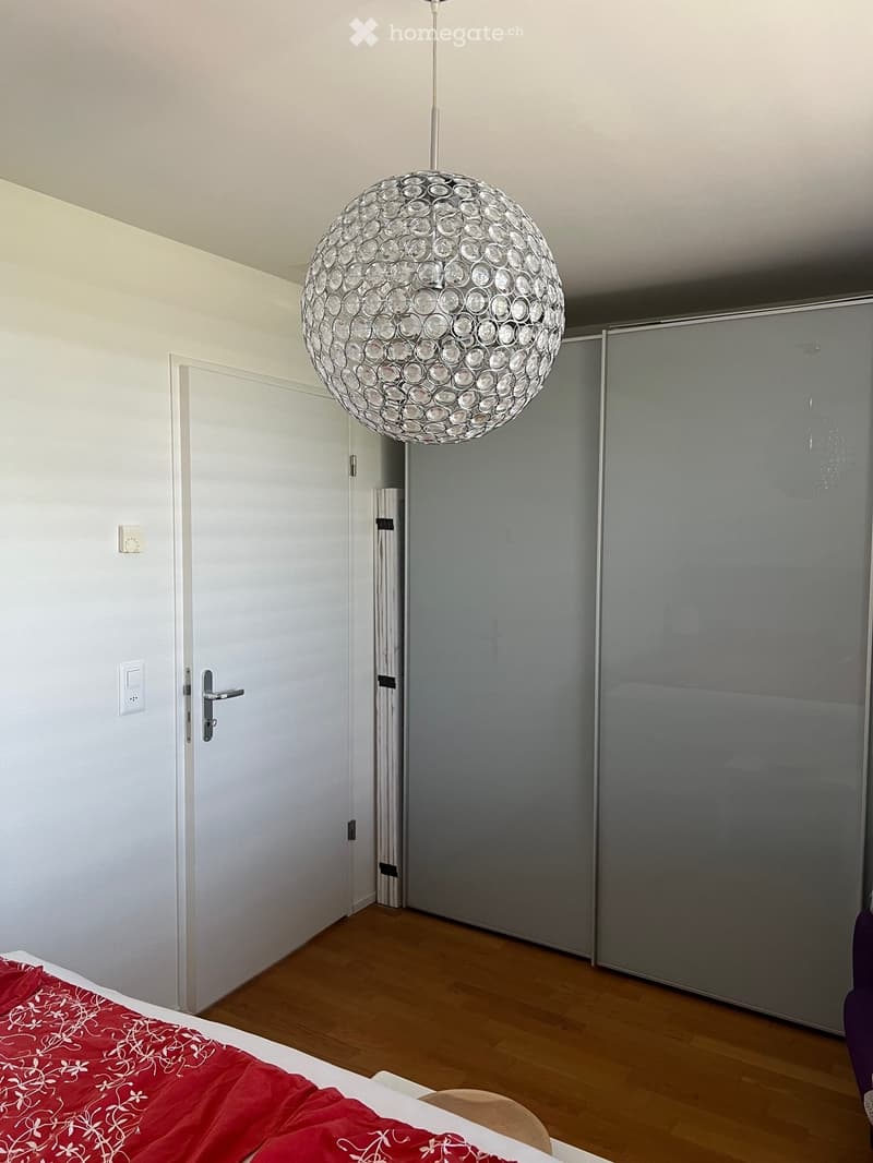 Modern bedroom with wooden flooring, a large spherical pendant light, a bed with red bedding, and a sliding wardrobe.