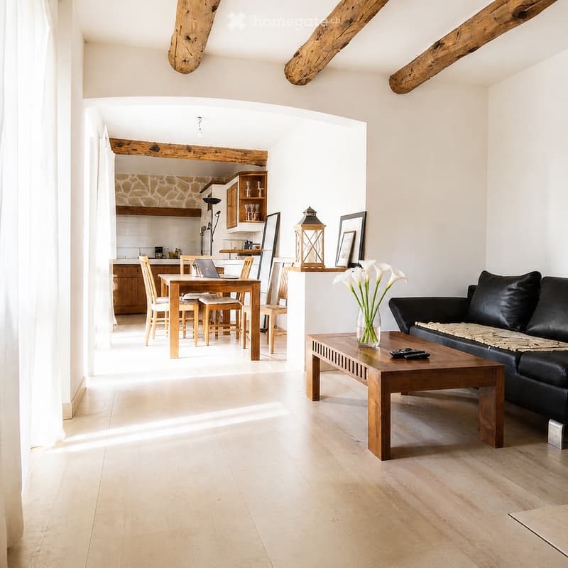 Open-plan living and dining area with exposed wooden beams, light wooden flooring, a black leather sofa, wooden coffee table, dining table with chairs, and a kitchen area in the background.