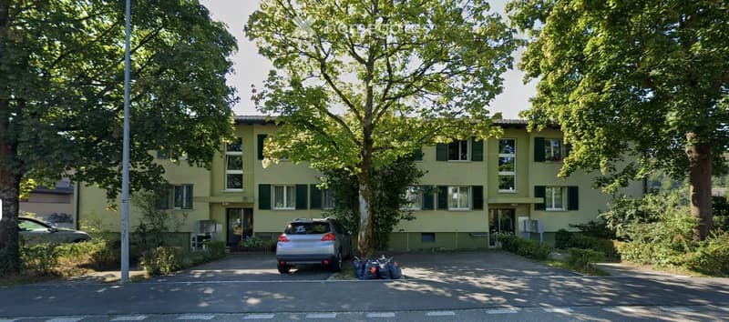 A two-story residential building with light yellow walls, green shutters, and multiple windows. A silver SUV is parked in the driveway, and there are trees and bushes surrounding the property.
