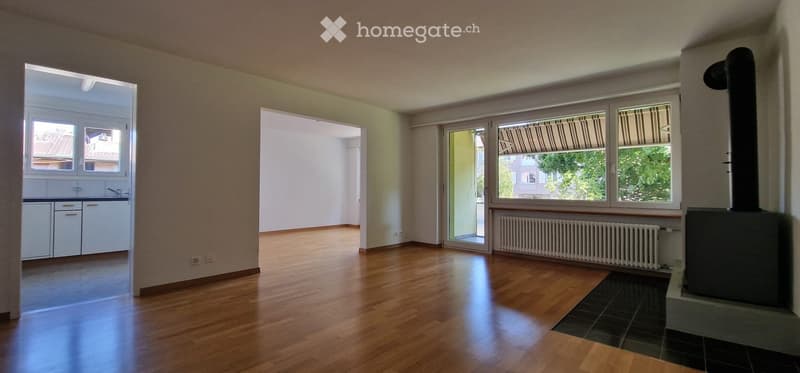 Empty room with wooden flooring, large windows, fireplace, and open doorways leading to other rooms.