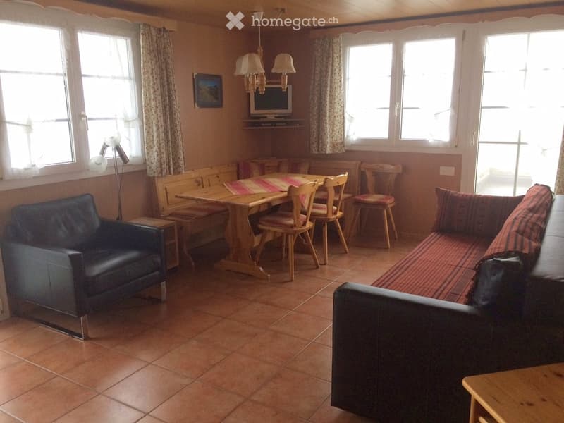 A cozy living room with a sofa, armchair, dining table, chairs, and a television. The room features wooden furniture, tiled flooring, and large windows with curtains.