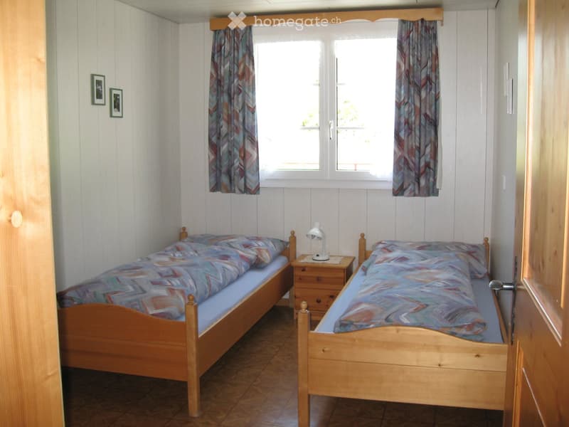 Cozy bedroom with two single wooden beds, matching bedding, a nightstand with a lamp, and framed pictures on the wall.