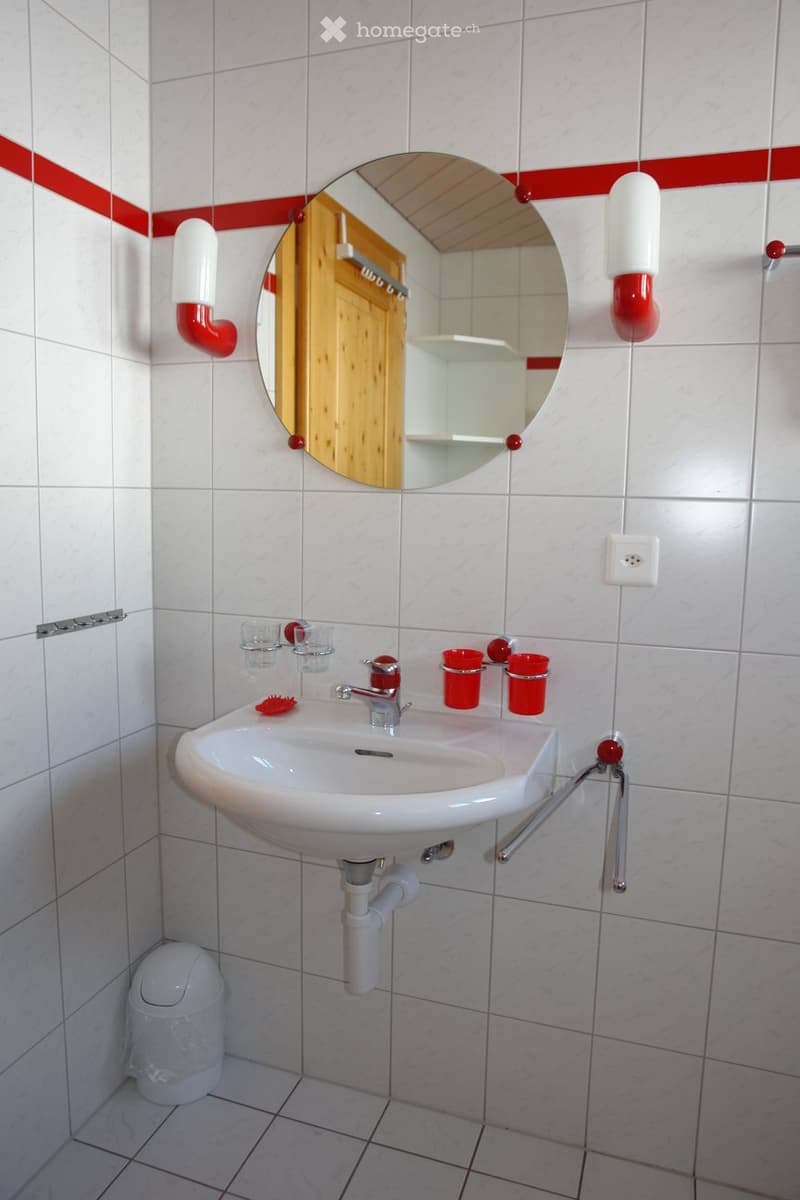 Bathroom with white tiled walls and floor, a white sink with a chrome faucet, a round mirror with a wooden frame, two red cups, a white toilet, and a soap dispenser.