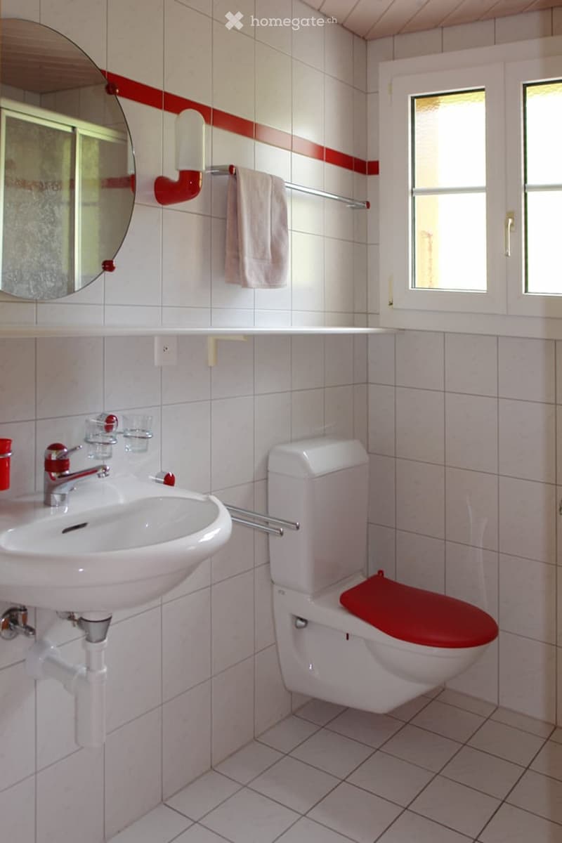 A small bathroom with white tiles, a red accent border, a white sink with a chrome faucet, a white toilet with a red seat cover, a round mirror, and a towel hanging on a red rack.