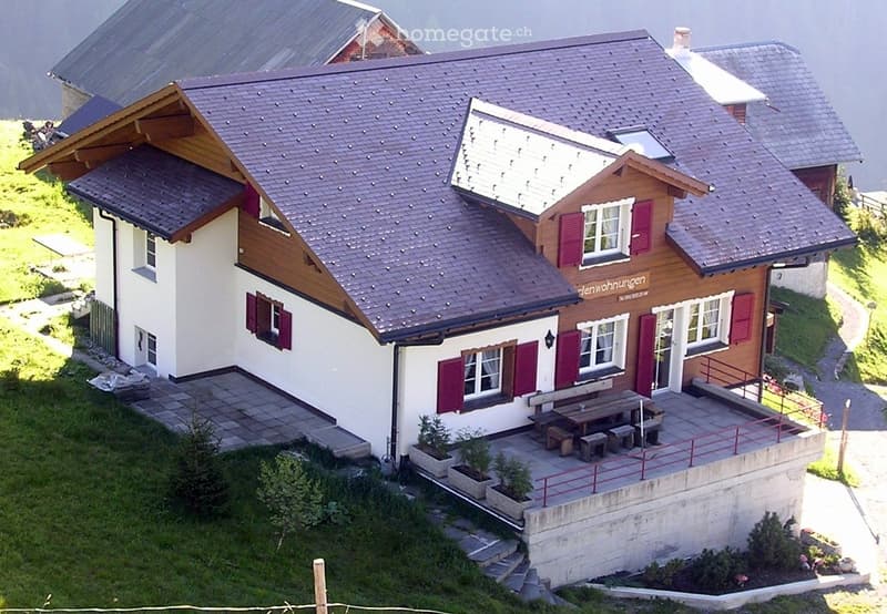 A two-story house with a gray tiled roof, white and brown exterior, red shutters, and a small balcony area with benches and potted plants.