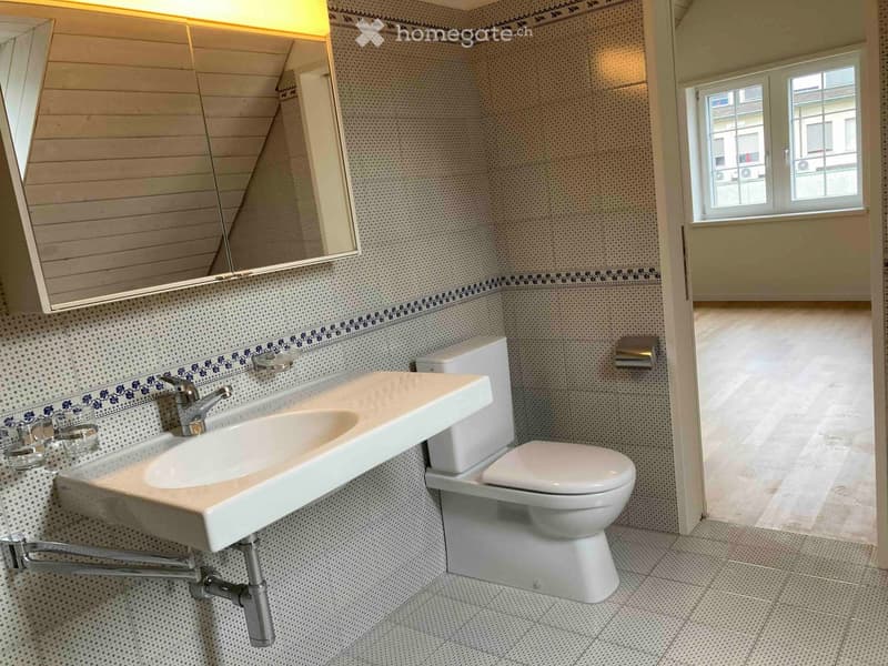 Bathroom with a white sink, toilet, and mirror. The walls are tiled with a decorative border, and there is a window letting in natural light.