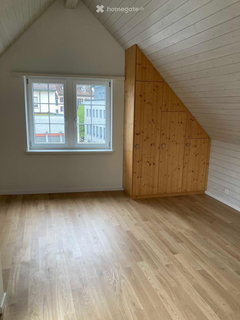 Empty attic room with wooden floors, slanted wooden ceiling, and a built-in wooden closet. A large window offers a view of neighboring buildings.