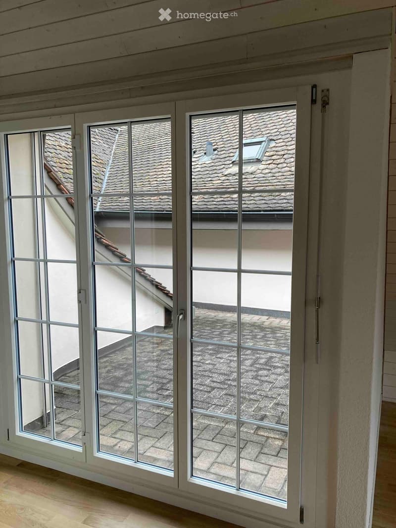 Empty room with large windows showing a view of a tiled roof and brick patio area. The room has wooden flooring and white walls.