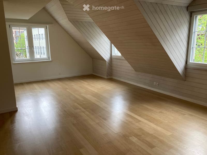 Empty attic room with wooden floors, slanted wooden ceiling, and large windows offering natural light.