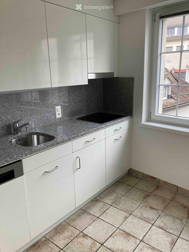 Modern kitchen with white cabinets, granite countertop, stainless steel sink, and built-in oven. Features tiled flooring and a window overlooking a rooftop.