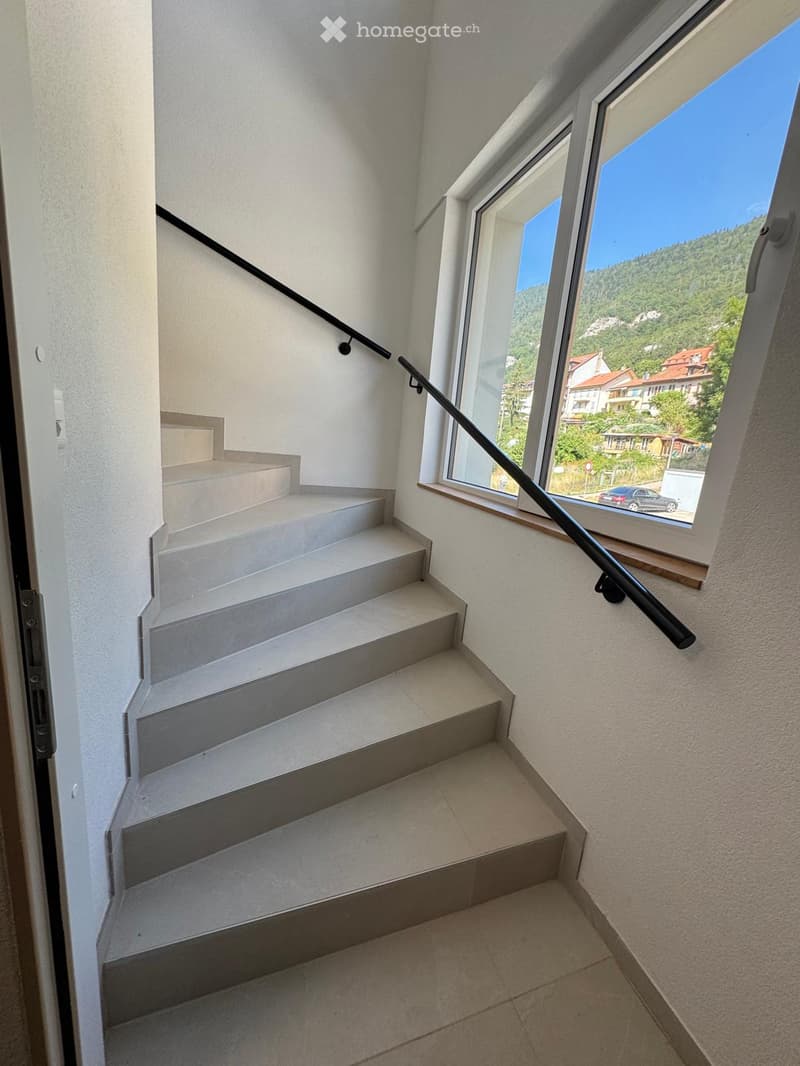 Modern staircase with white walls, grey steps, and a black handrail, leading to a view of a town and landscape through a large window.
