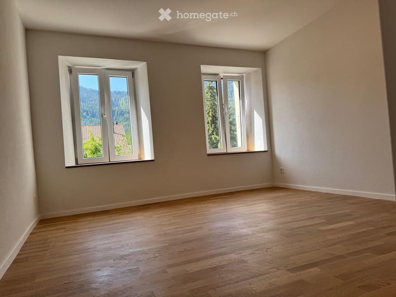 Empty room with wooden flooring and two large windows offering a view of greenery and a roof outside.