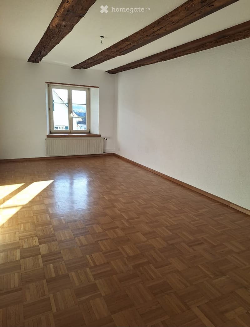 Empty room with wooden herringbone flooring, white walls, exposed wooden beams on the ceiling, and a single window with a radiator below it.