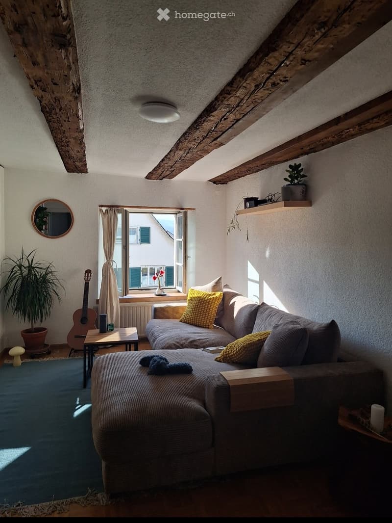 Cozy living room with exposed wooden beams, a large window, a grey sectional sofa with yellow pillows, a coffee table, a guitar, and potted plants.