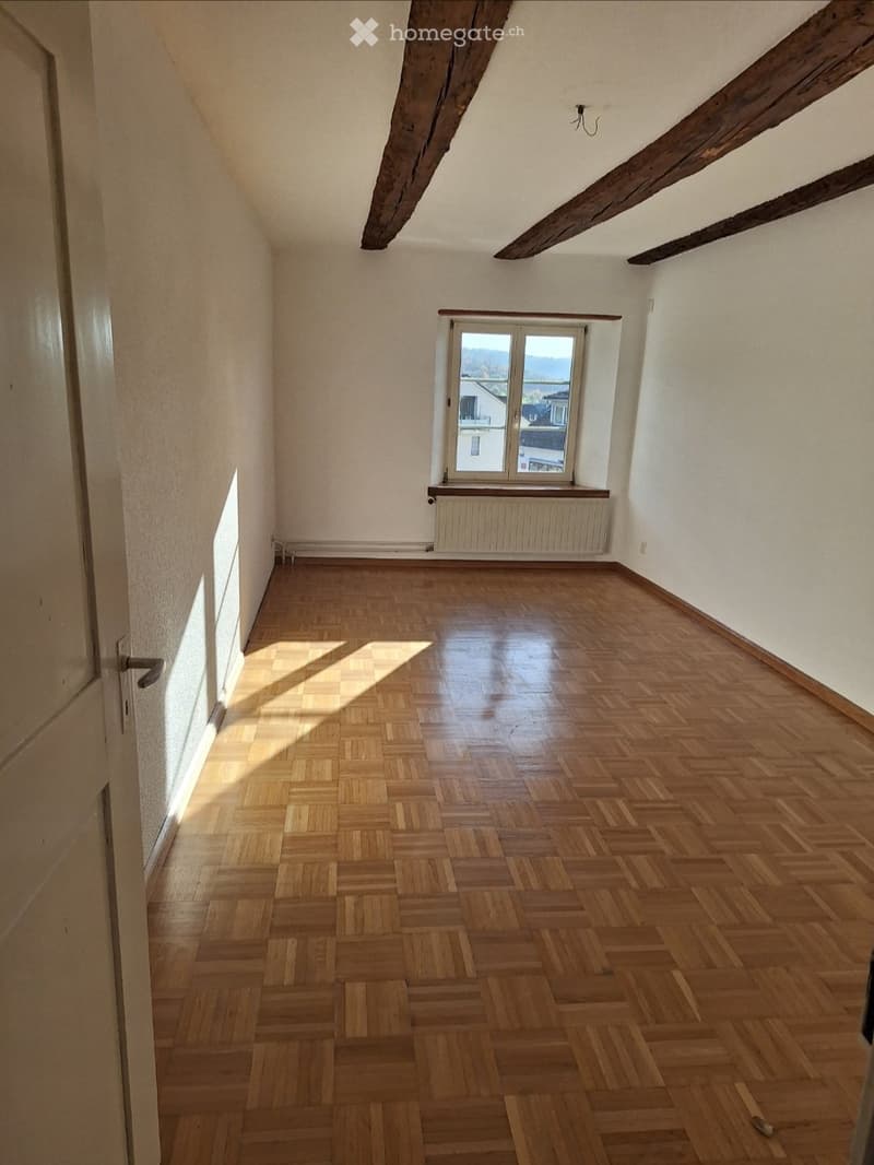 Empty room with wooden parquet flooring, white walls, exposed wooden beams on the ceiling, and a single window with a radiator below it.