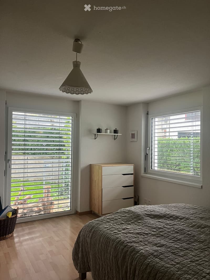 Modern bedroom with a large bed, wooden chest of drawers, wicker basket, wall-mounted shelf, and a hanging lamp. Large windows with blinds and a sliding glass door lead to a green outdoor area.