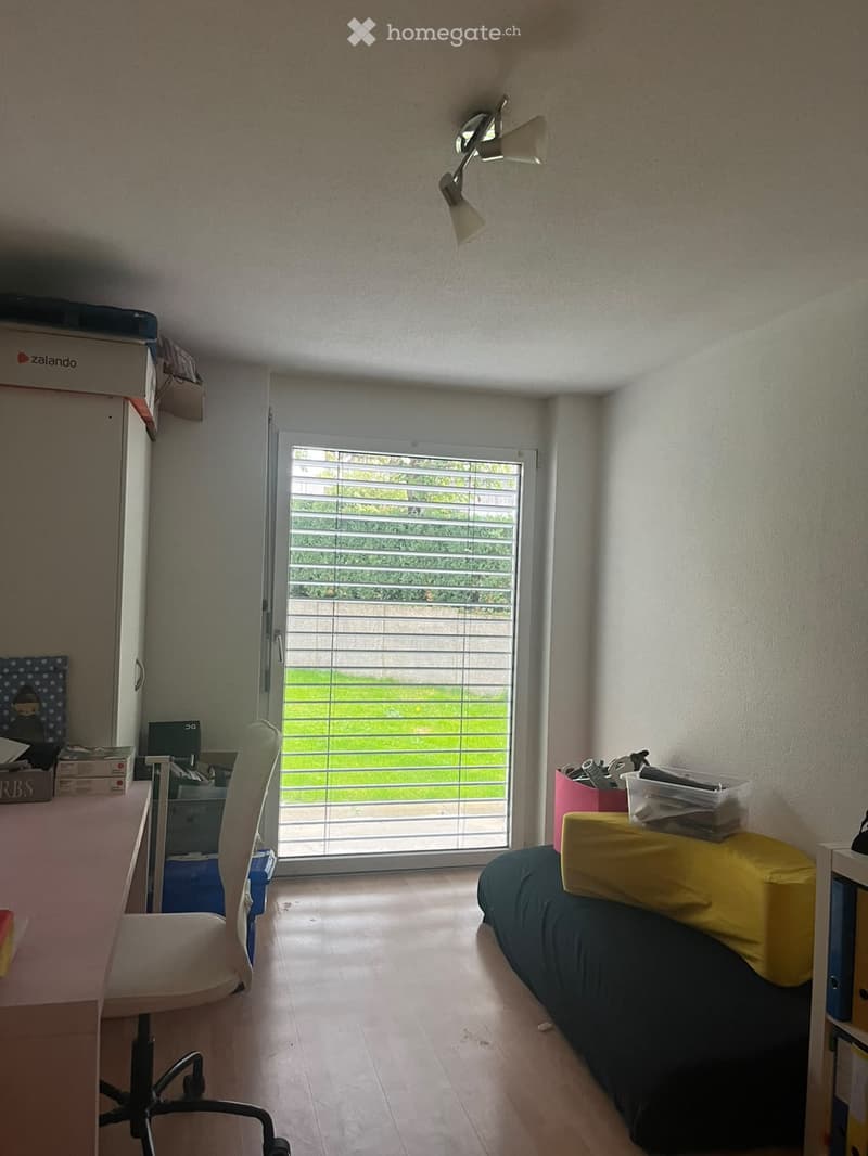 A furnished room with a bed, desk, chair, and storage boxes. The room has a large window with blinds, allowing natural light to enter.