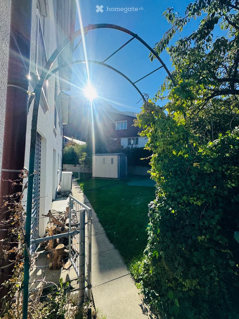 A narrow garden pathway lined with greenery, leading to a small shed and a house in the background. The sun shines brightly, creating lens flare effects.