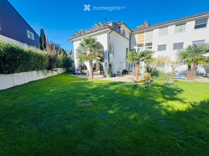 Well-maintained garden with green lawn, palm trees, and a white house with arched windows and a balcony.