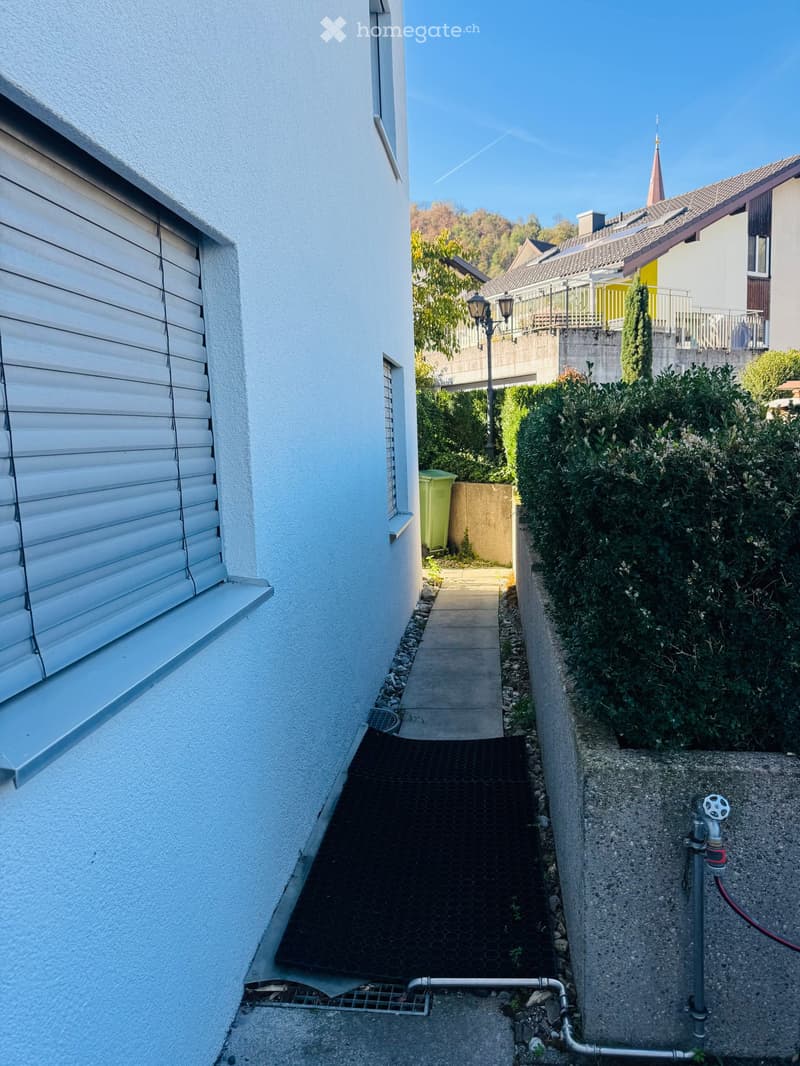Narrow pathway between two buildings with a small garden area, concrete walls, and a water tap.