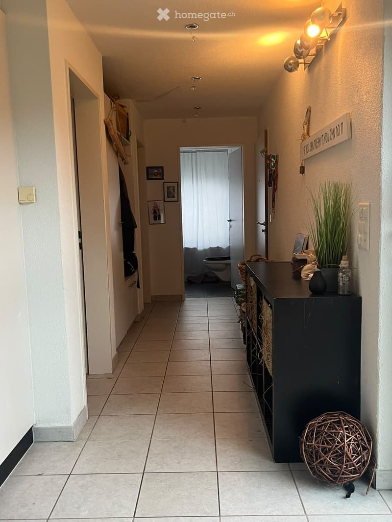 A well-lit hallway with tiled flooring, white walls, and several doors leading to different rooms. There is a small table with decorative items, a coat rack, and framed pictures on the walls.