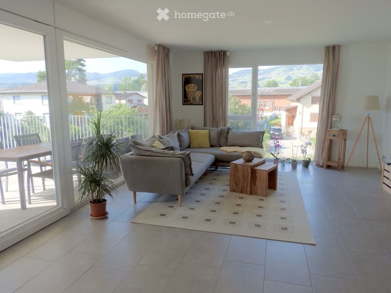 Bright living room with large windows offering a scenic mountain view, featuring a grey sectional sofa, wooden coffee table, side table, potted plants, and a dining area with a table and chairs.