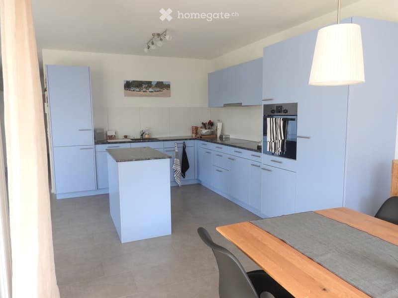 Modern kitchen with light blue cabinets, central island, sink, oven, and dining area with table and chairs.