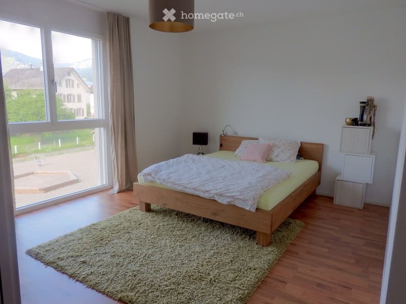 Bright bedroom with wooden floor, large window with view, bed with pillows and blanket, shaggy rug, wall-mounted lamp, and small nightstand with mirror.
