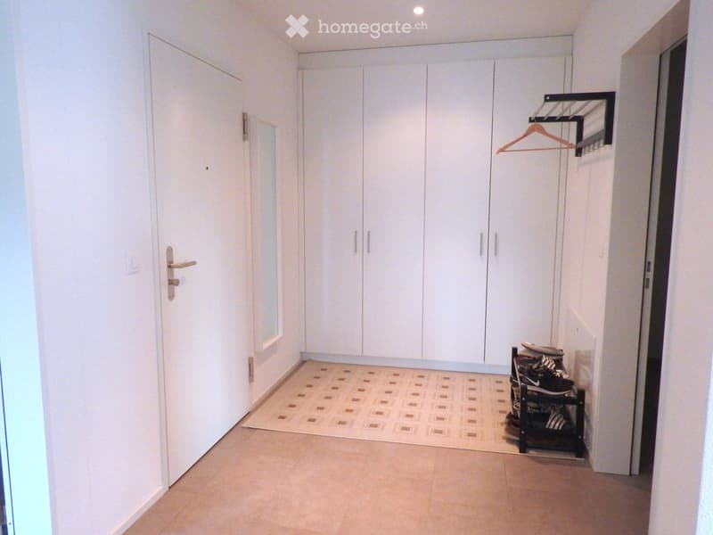 A clean, well-lit hallway with white walls, a tiled floor, built-in white wardrobes, a coat rack with a single hanger, and a small shoe rack holding several pairs of shoes.