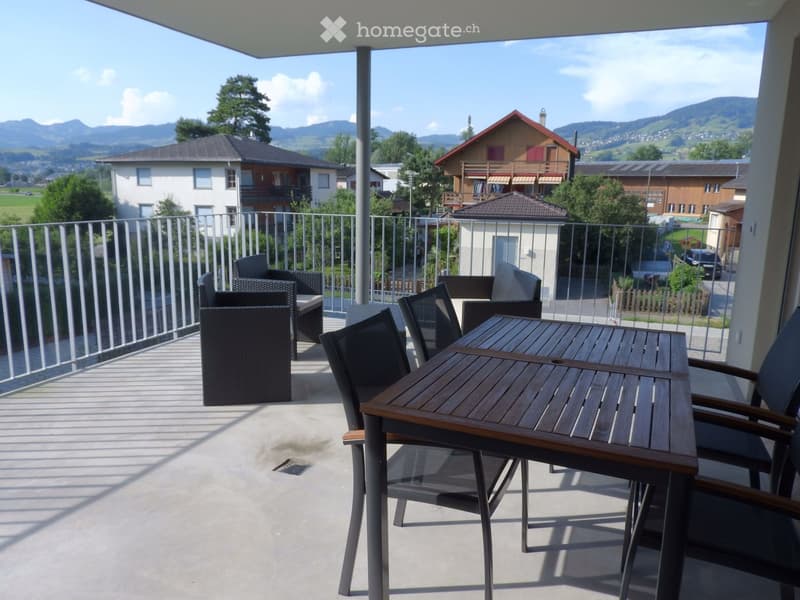 Balcony with a wooden table and several chairs, overlooking a scenic mountain view with houses and greenery.