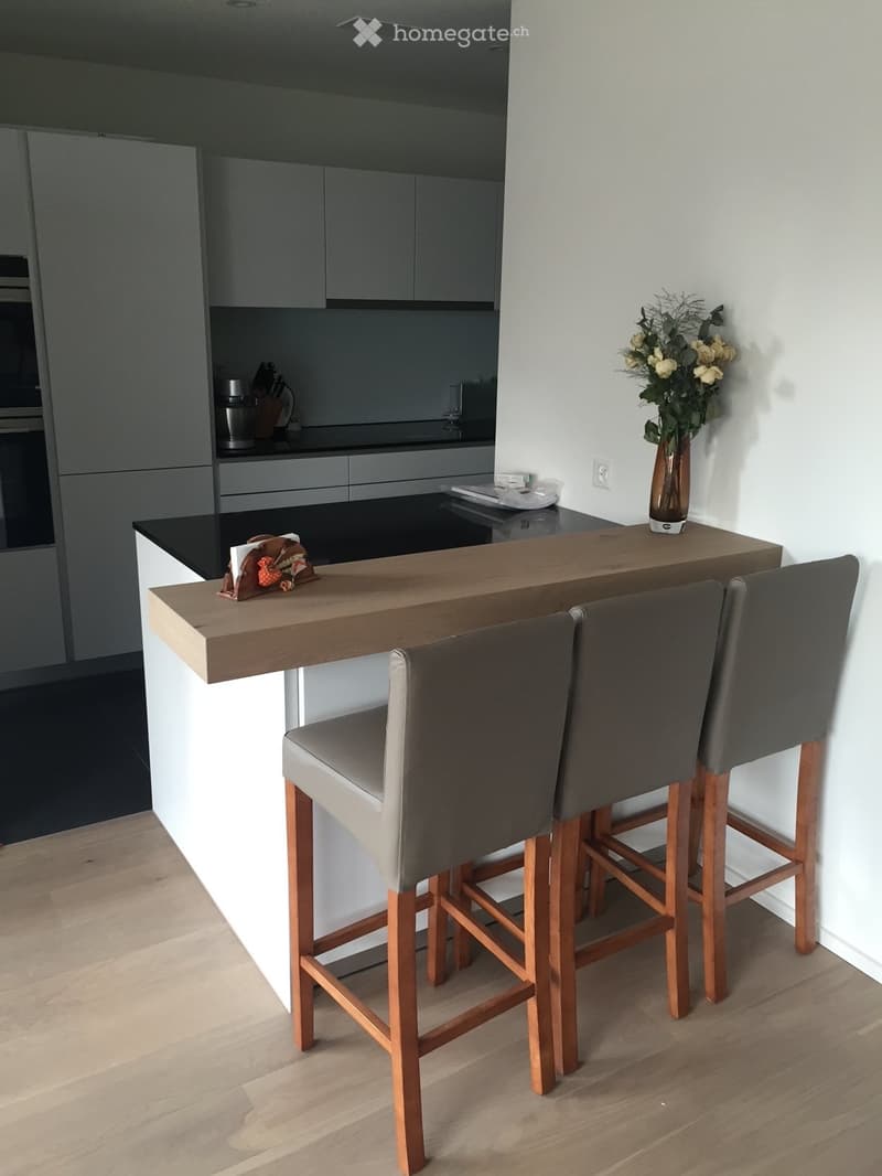 Modern kitchen with grey cabinets, black countertops, a wooden bar table, three grey bar stools, and a vase with flowers on the counter.