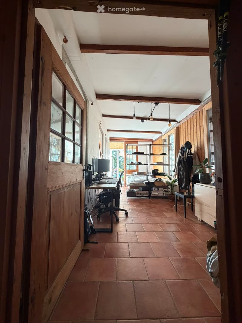 A spacious, well-lit room with wooden beams and terracotta tiles, featuring a desk with a computer, a shelf with books and items, a small stool, and a potted plant.