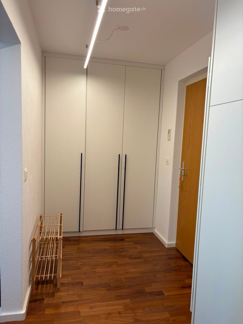 Empty hallway with wooden flooring, white walls, and a large white closet with double doors. A small wooden shelf is placed against the left wall.