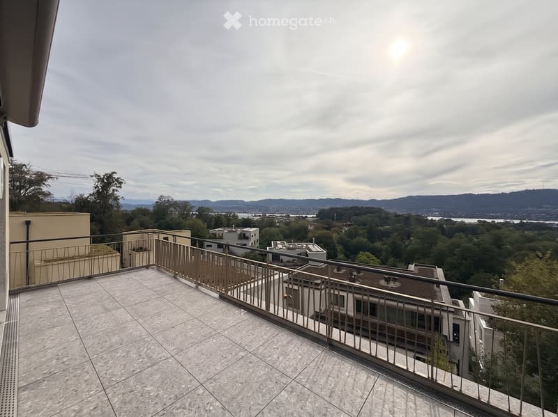 Balcony with tiled floor and metal railing, overlooking a cityscape with trees and distant mountains.