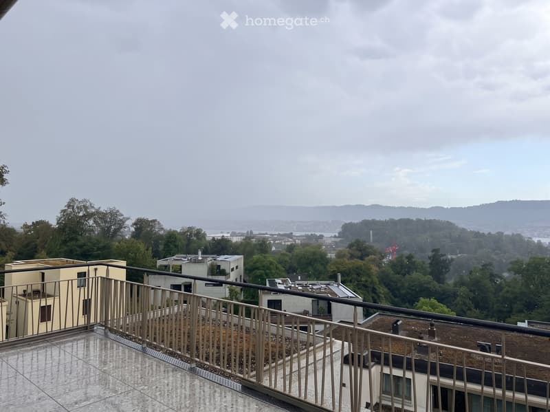 Balcony with metal railing, tiled floor, overlooking a cityscape with trees and buildings.