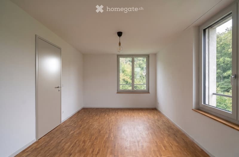 Empty room with wooden flooring, white walls, and large windows offering a view of greenery outside.