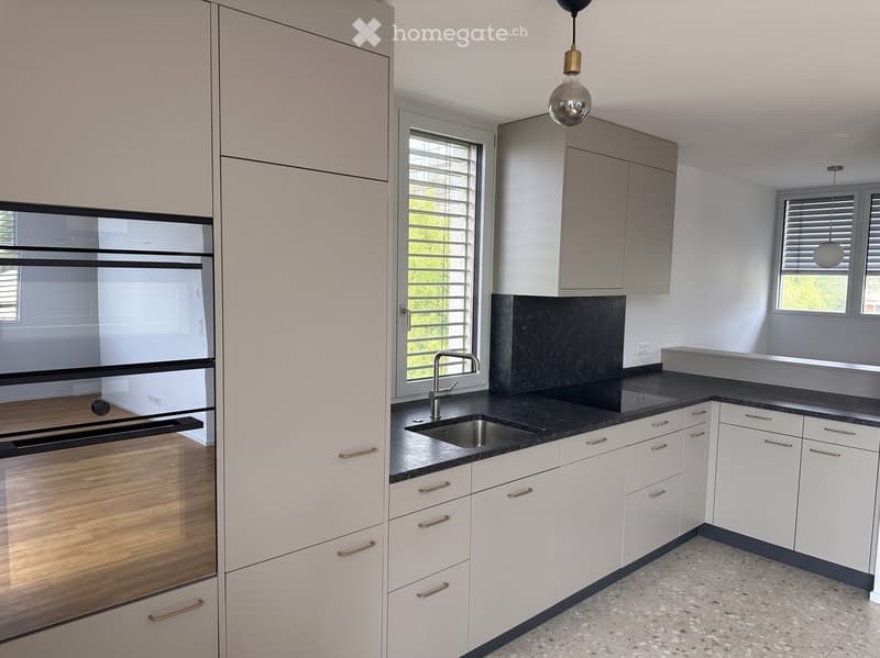 Modern kitchen with white cabinets, black countertops, a stainless steel sink, and a built-in oven. Features include a pendant light and large windows with blinds.