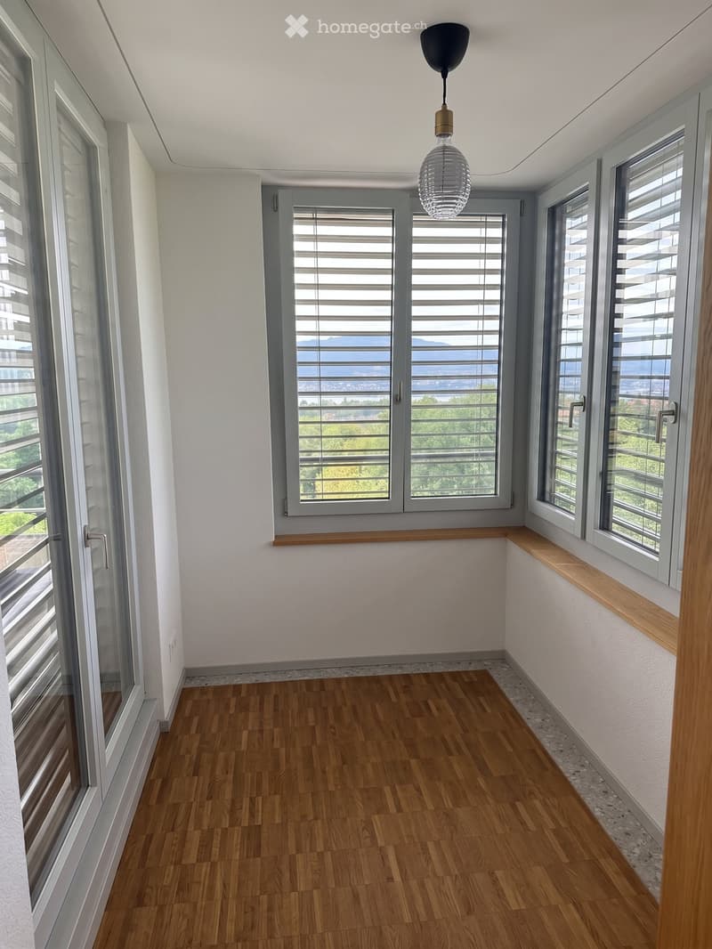 Empty room with wooden flooring, large windows with blinds, and a hanging light fixture.