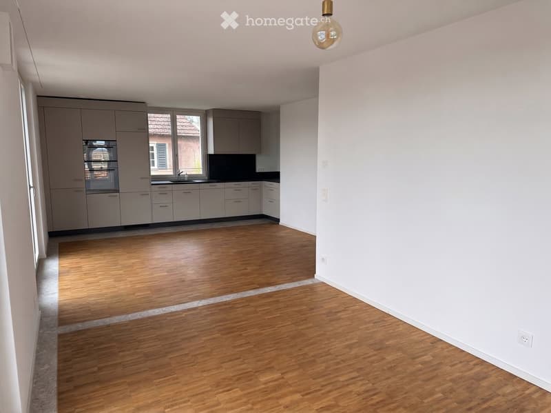 Empty room with light wooden flooring, white walls, and a small kitchen area with cabinets and a sink visible in the background.