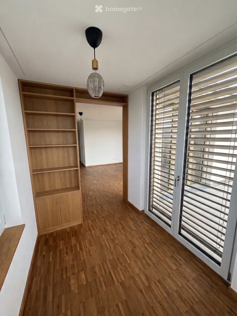 Empty room with wooden flooring, white walls, large windows with blinds, and a built-in wooden shelving unit.