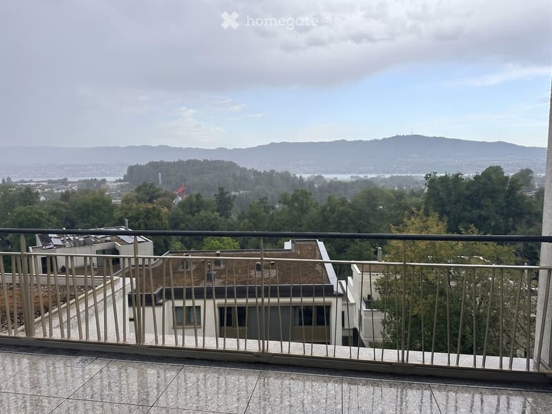 Balcony with metal railing, tiled floor, overlooking a cityscape with trees, buildings, and distant hills.