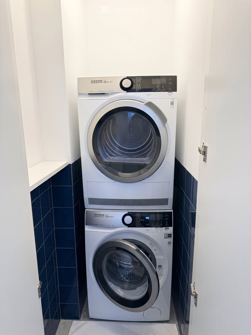 Modern bathroom with a white washing machine and dryer stacked vertically, featuring a sleek digital control panel and a dark blue tiled wall.